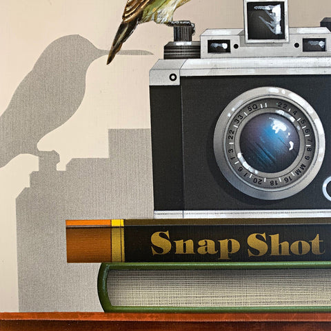Detail of Painting of a ruby-throated hummingbird perched atop a vintage camera on two books, one titled Snapshot, on a tabletop by James Carter at Cottage Curator - Sperryville VA Art Gallery